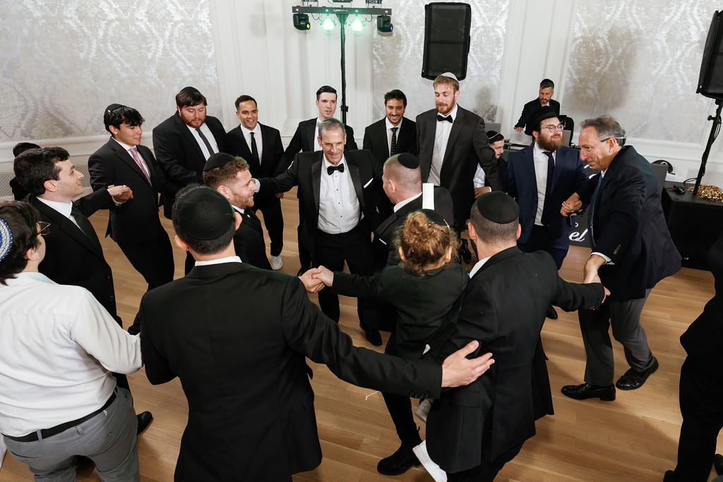 Groom in a black suit surrounded by men in formal attire during a dance and celebration indoors, with an upbeat crowd forming a tight circle.