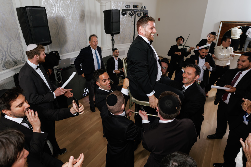 The groom laughing and greeting guests during a lively wedding moment, surrounded by men clapping and holding lighted sticks in a ballroom setting.