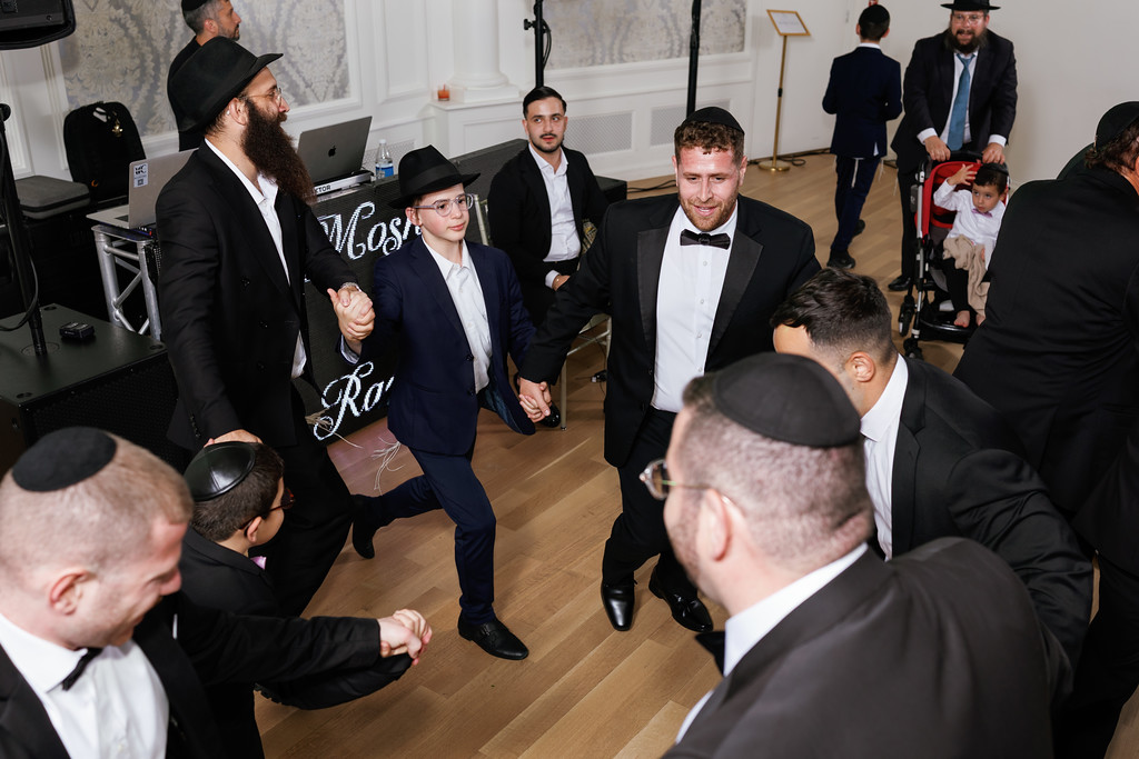 A groom smiles while moving through a lively group of guests on the dance floor during a Jewish wedding celebration, with a woman in a cap and a rabbi visible nearby.
