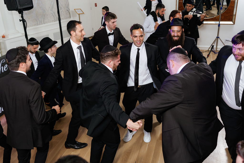 At a Jewish wedding celebration, the groom in a black suit and kippah shares a joyful handshake with a friend as guests in dark suits gather around on the dance floor.