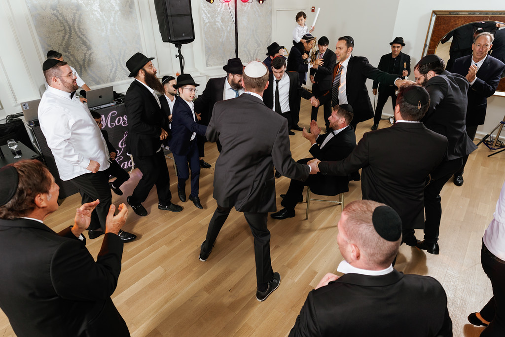 A group of men in black hats and suits dance together indoors while other guests clap and watch, creating an energetic wedding celebration moment.