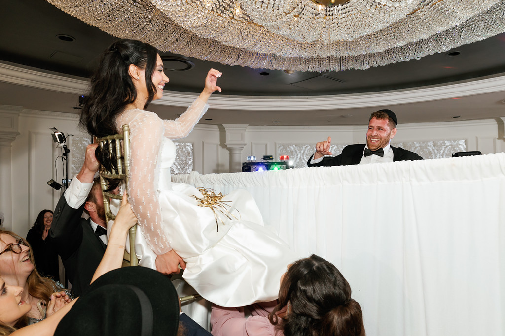 Bride dancing joyfully with her hands raised while the groom smiles behind a white draped setup; guests watch and celebrate at the reception.