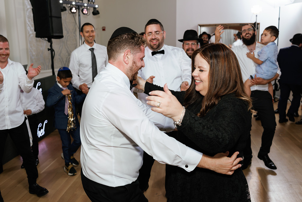 A couple dances in an indoor celebration, hugging and smiling while guests and children watch in the background.