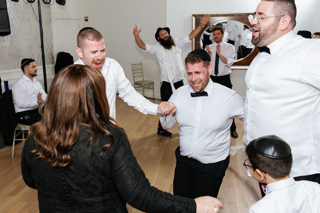 Frum wedding celebration dance scene: the groom in a white shirt and black bow tie laughs while holding hands, with guests cheering in the background.