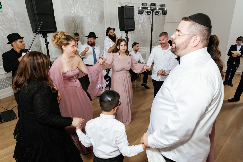 A woman in a mauve dress dances at a wedding reception while guests in the hall smile and enjoy the moment, seen from a candid side angle with a rabbi in the foreground.