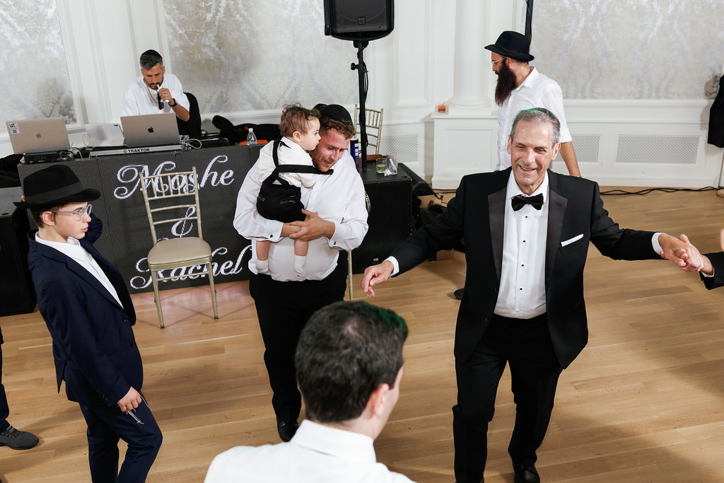A father in a white shirt holds his toddler in front of a DJ setup while a man in a tuxedo smiles during a Jewish simcha celebration.