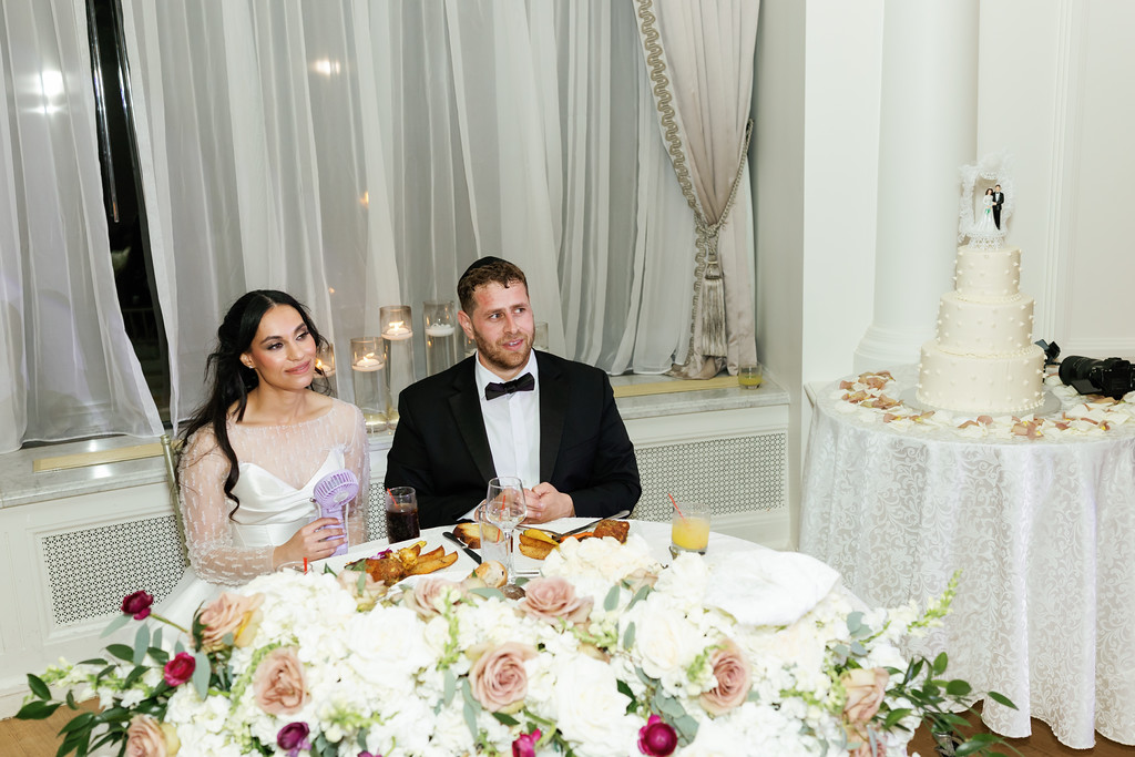 Frum bride and groom seated at a decorated reception table with floral centerpieces, smiling while dining in front of wedding drapery and a cake in the background.