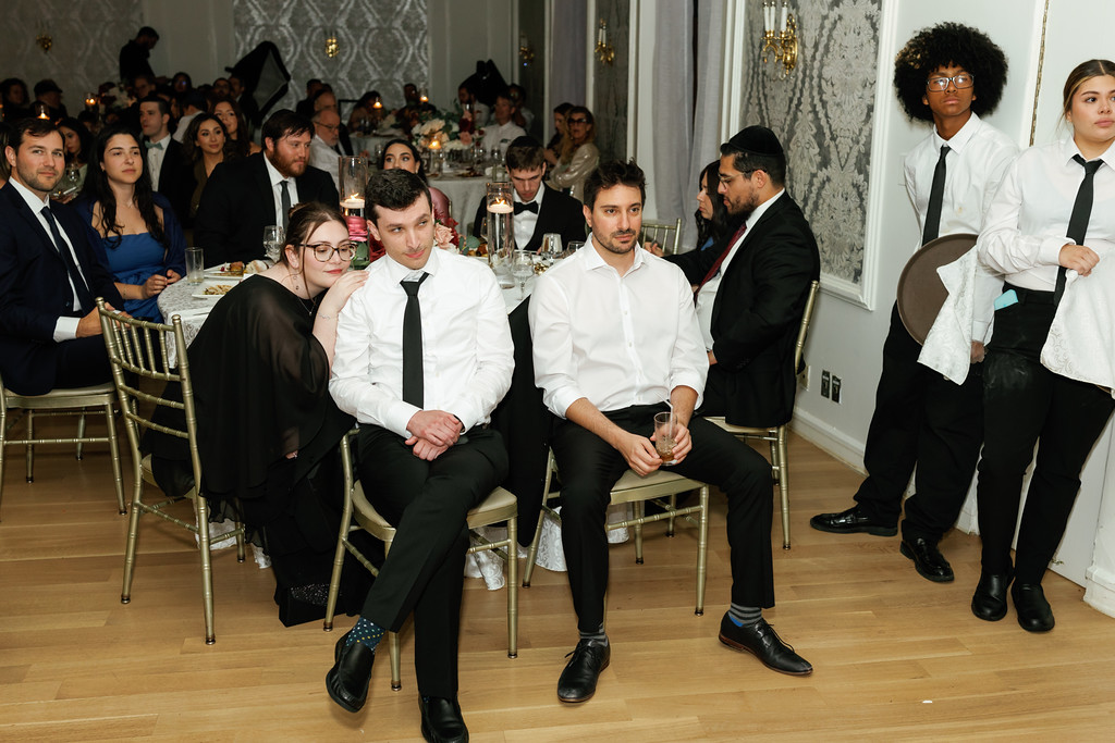Guests seated at a reception table during a celebration, with a man in a white shirt holding a drink in the foreground and other guests in formal attire in the background.