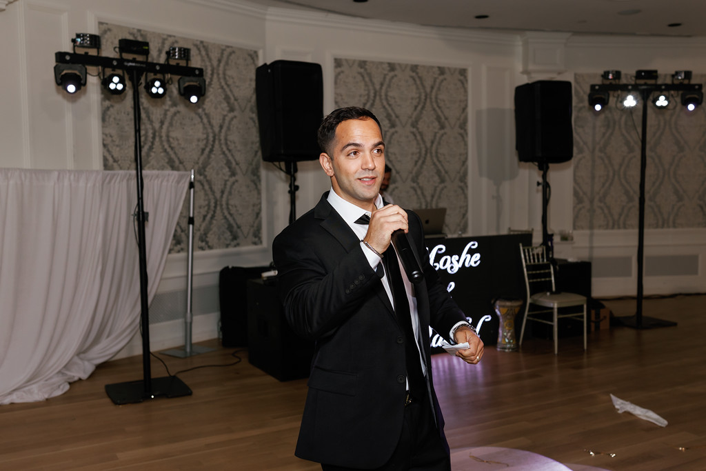 Smiling groom/MC in a black suit holding a microphone while giving a speech in an indoor event hall with stage lighting and speakers in the background.