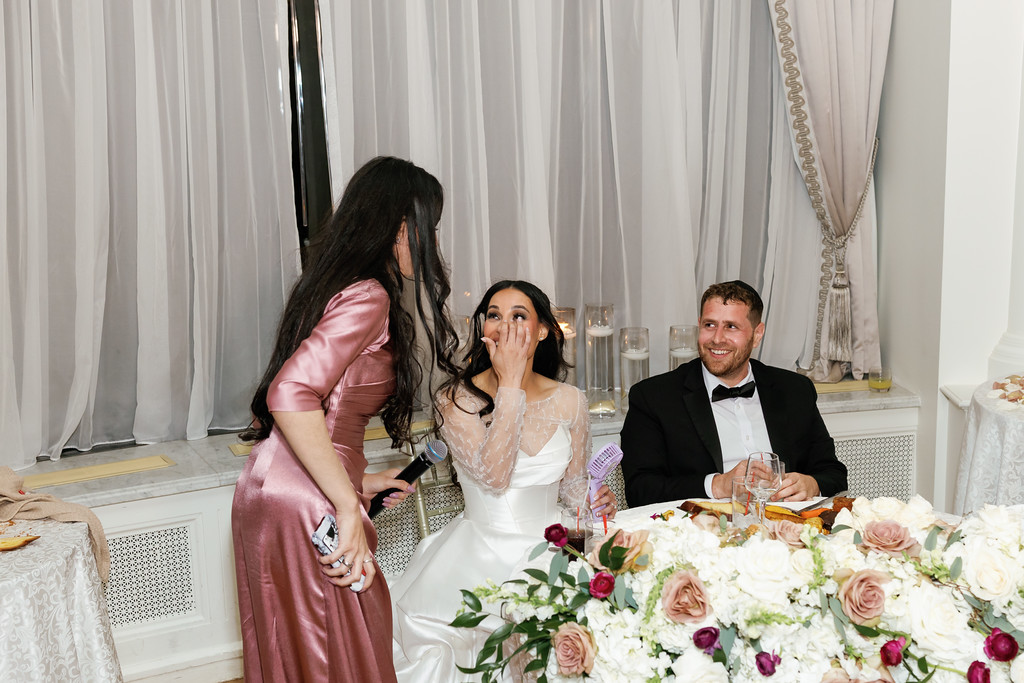At a wedding reception table, the bride in a white gown covers her mouth while laughing, smiling next to the groom in a black tuxedo; a guest in a pink dress leans in as they celebrate.