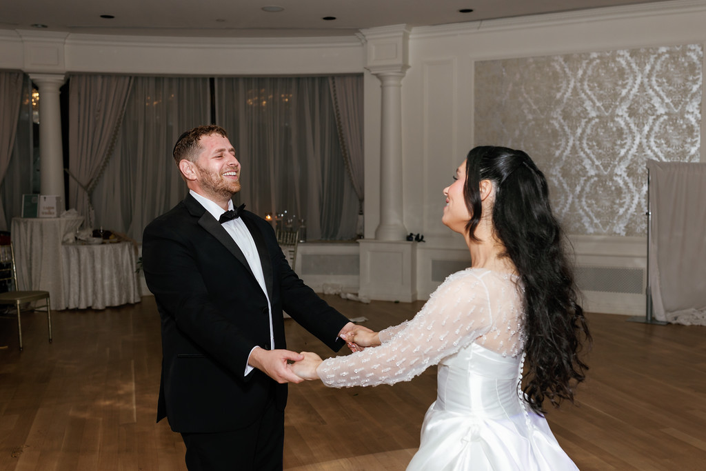 A bride and groom holding hands while dancing at their wedding reception, smiling at each other in a ballroom with draped curtains and patterned lighting.