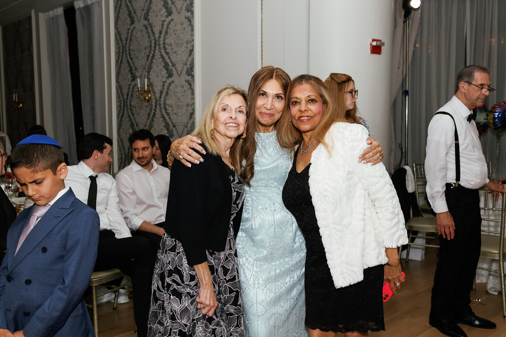 Three women pose closely together in a warm embrace at a wedding reception, smiling in front of a softly lit event hall.