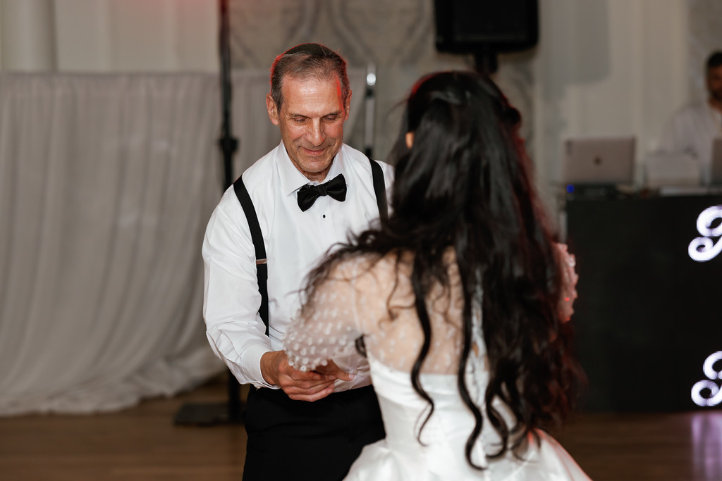 A groom in a black bow tie and suspenders smiles while looking down at the bride in a white gown, holding her hands during the wedding celebration.