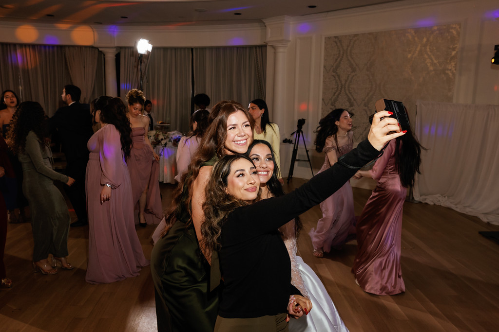 Smiling bride and guests dancing at a wedding reception, with colorful purple uplighting and a phone held up in the background.