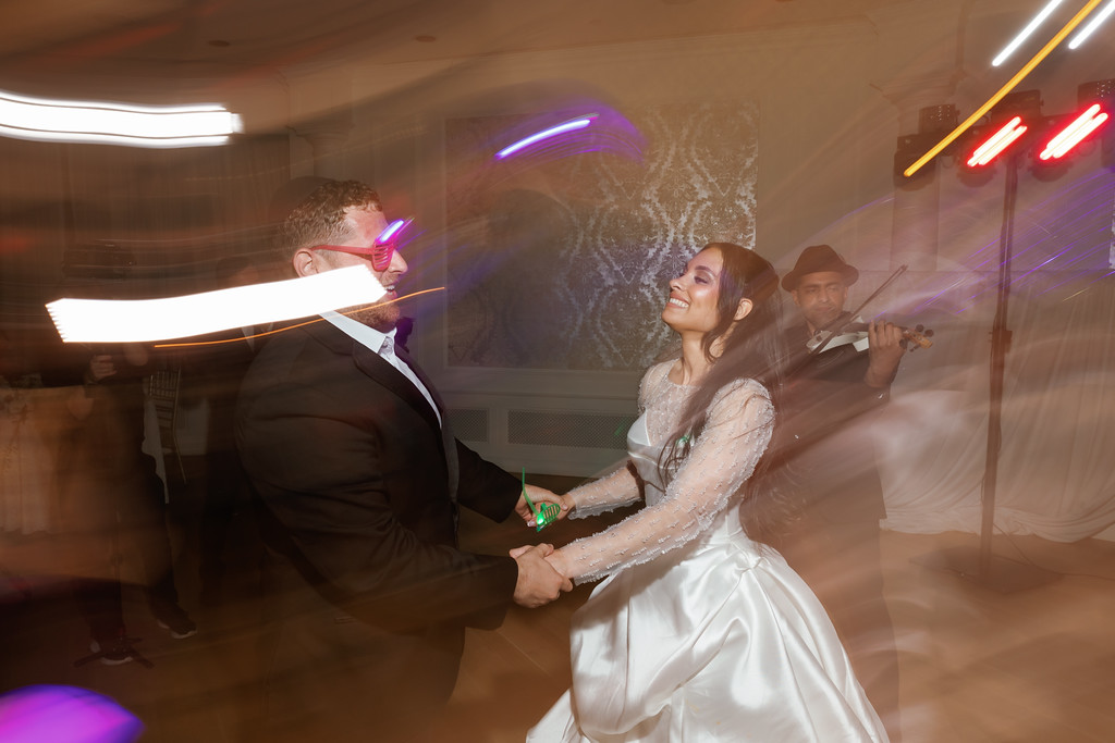 Bride and groom smiling while dancing, holding hands during the reception as party lights create light trails in the background.