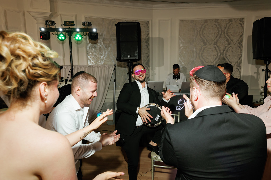 Guests dance in a circle at a wedding reception as the groom and party members smile and clap in the center under event lights.