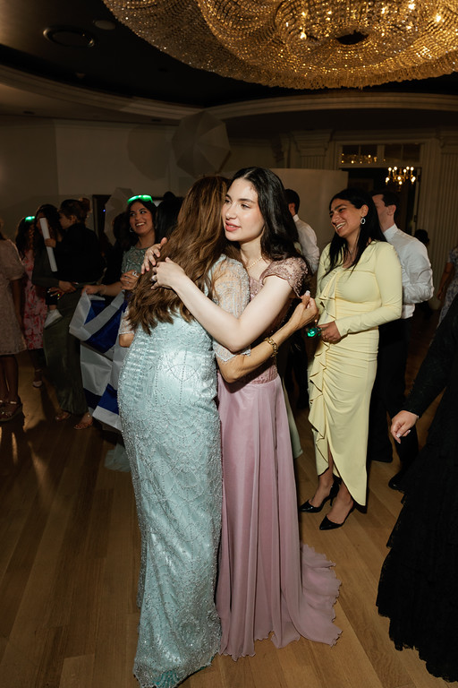 Bride and guests dancing at a wedding/banquet, the bride shown in three-quarter profile smiling while another woman embraces her from the side.