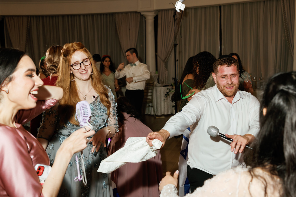A smiling couple dances at a wedding reception with a microphone while guests cheer in the background.