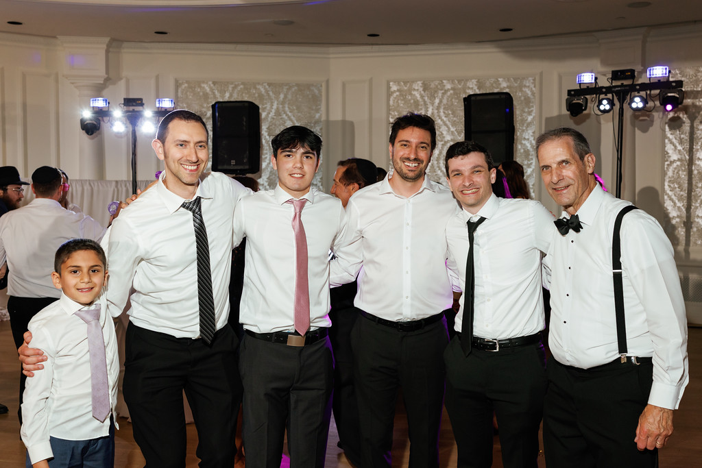 Group photo of a smiling adult man and a young boy standing with several men in white shirts and black ties at a wedding reception.