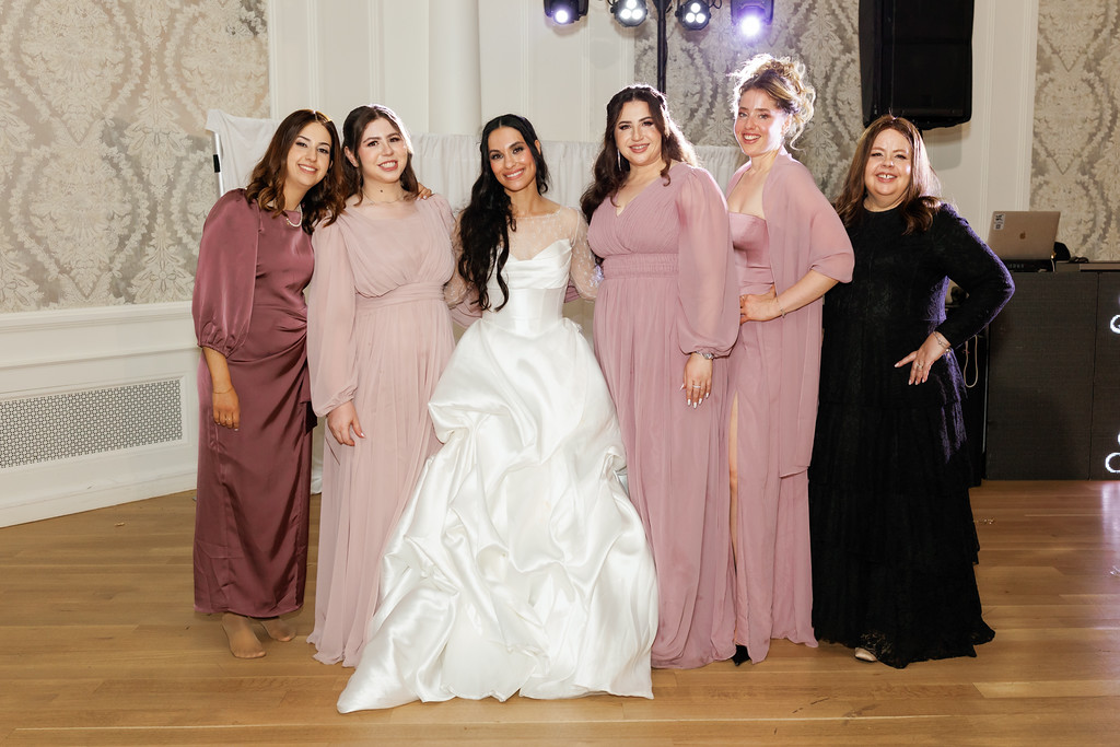 Bride in a white gown posing center with five bridesmaids in mauve and black dresses at a reception hall, all smiling toward the camera.