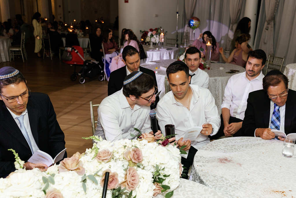Groom and family seated at a reception table looking down at papers during a formal moment, with floral centerpieces in the foreground.