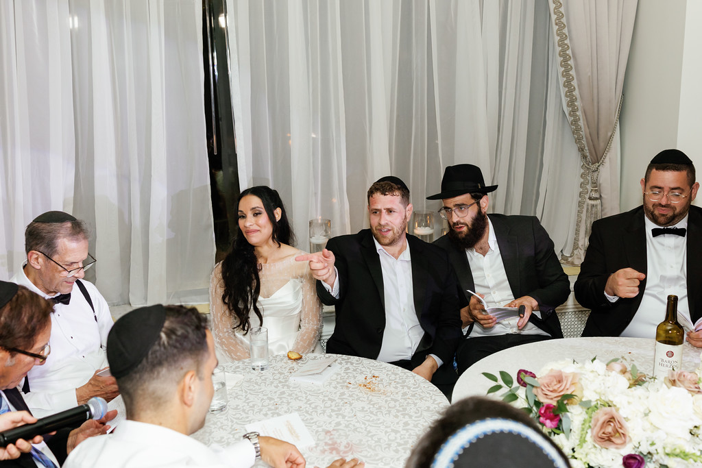 Bride and groom seated at a wedding reception table with family and guests around them, the groom gesturing while others smile and talk.