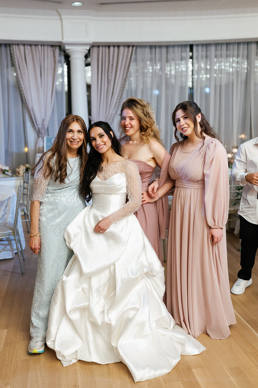 Frum wedding celebration with the bride in a white dress and friends/family standing close together and smiling in a decorated event hall.