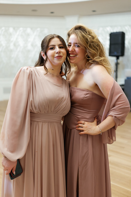 Two women in blush dresses smiling as they embrace and pose together indoors during an event.