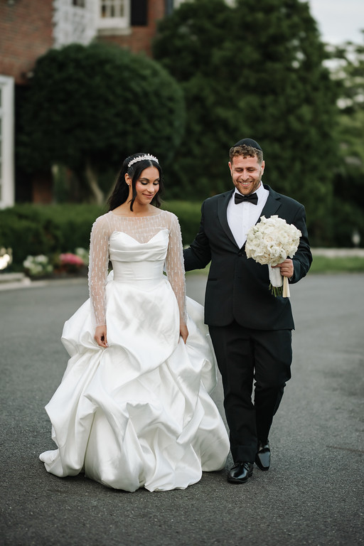 Bride in a white lace wedding dress and groom in a black tuxedo walking together outdoors on a garden walkway, looking down as they move forward.