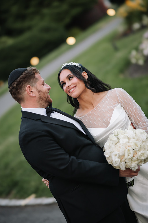 A groom in a black tuxedo holds his bride close as she smiles in a white bridal dress while holding a bouquet outdoors at night.