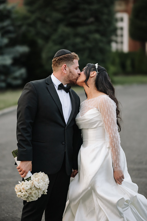 Frum wedding couple kissing outdoors in formal attire, with the bride in a white gown and sheer sleeves and the groom in a black tuxedo holding a bouquet.