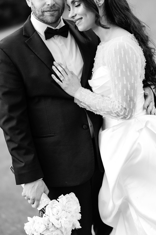 Black-and-white wedding portrait of a groom in a tuxedo and bow tie holding the bride close as they smile, with the bride’s hand on his chest and her bouquet in the lower frame.