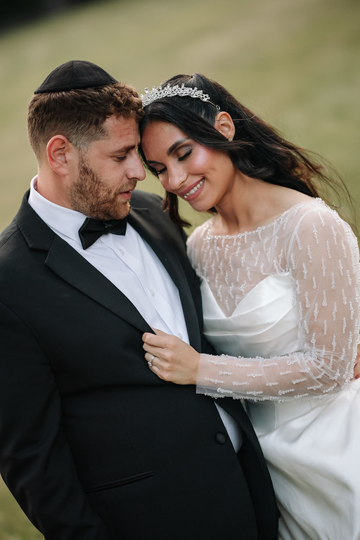Frum bride and groom in close embrace outdoors, smiling as the groom rests his hand on her arm and she leans into him.