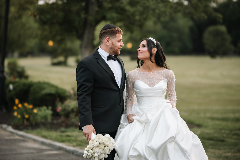 A bride in a white gown and veil and a groom in a black suit stand outdoors holding hands and looking at each other during a wedding moment.