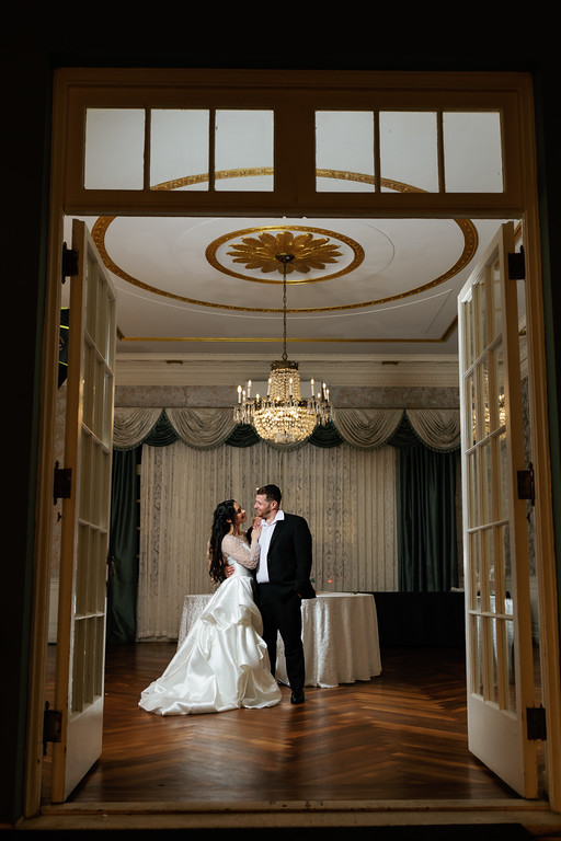 Bride and groom sharing an intimate moment facing each other inside an ornate doorway, with chandeliers and draped chuppah-style décor behind them.