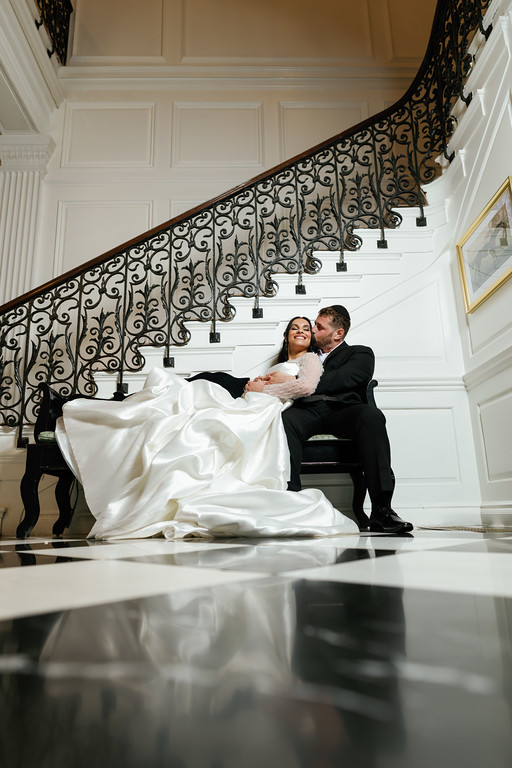 Bride and groom seated in an embrace, smiling at each other inside a grand white venue staircase with wrought-iron railing behind them.