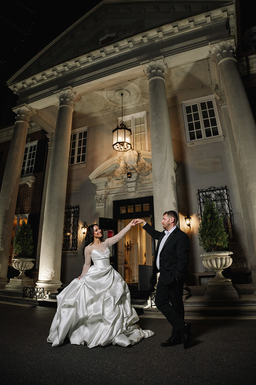 Framed by tall columns at a nighttime venue, a bride in a white gown and a groom in a black suit hold hands and face each other with warm smiles.