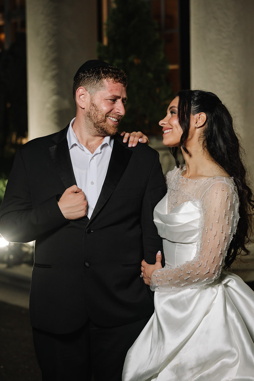 A smiling groom in a black suit and a bride in a white embroidered gown stand close together during the wedding, looking at each other.