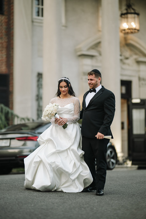 Frum bride in a white bridal gown holding a bouquet walks with her groom in a black suit and kippah outside a building, smiling as they move down the street.