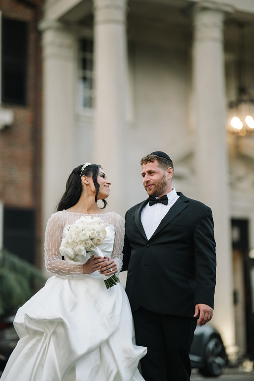 Frum bride in a white gown holding a bouquet, walking beside her groom in a black tuxedo outside during the wedding evening.