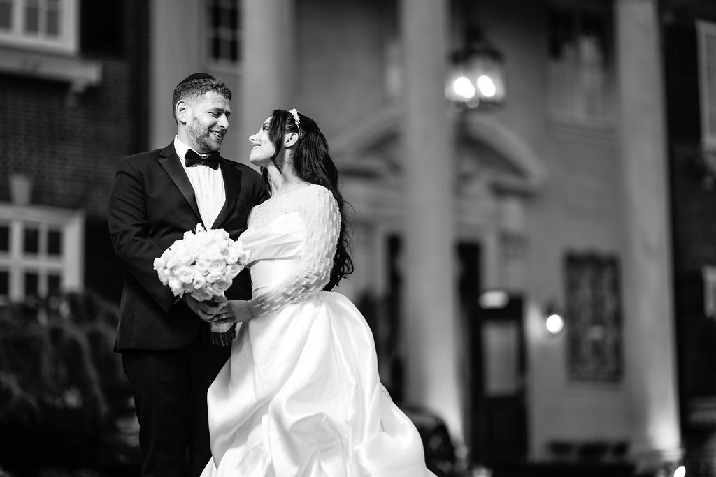 Black-and-white wedding photo of the bride and groom smiling at each other while holding a bouquet during the ceremony, with blurred lights in the background.