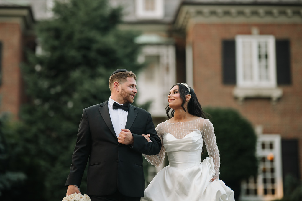 Groom in a black tuxedo and bow tie and bride in a white lace wedding dress stand outdoors smiling at each other, surrounded by greenery and a brick building.