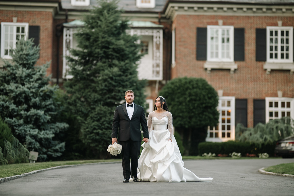 Bride and groom walking hand-in-hand on a garden driveway in front of a brick building, with the bride’s white gown flowing as they move toward the camera.