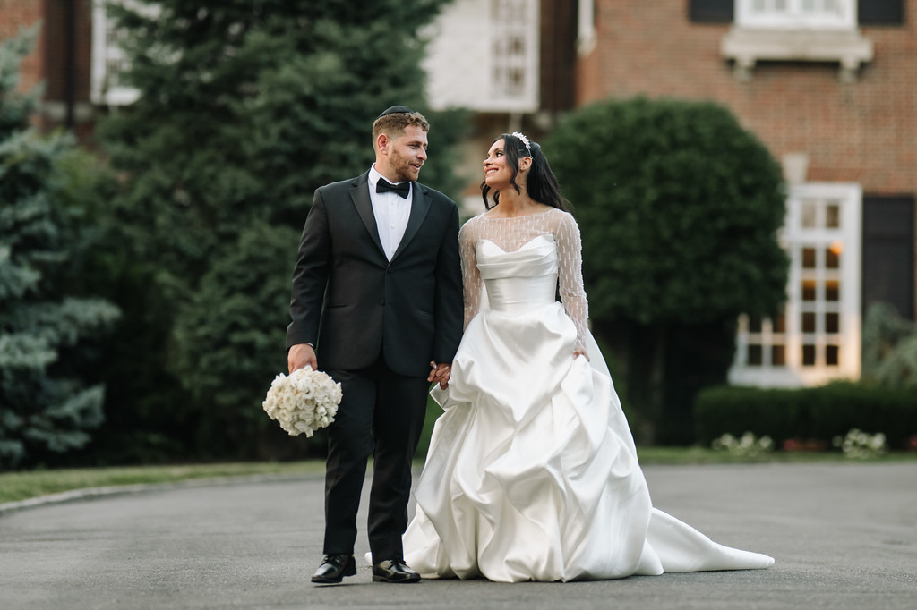 Bride and groom holding hands and gazing at each other outdoors on their wedding day, with the bride’s white gown flowing and a bouquet in the groom’s hand.
