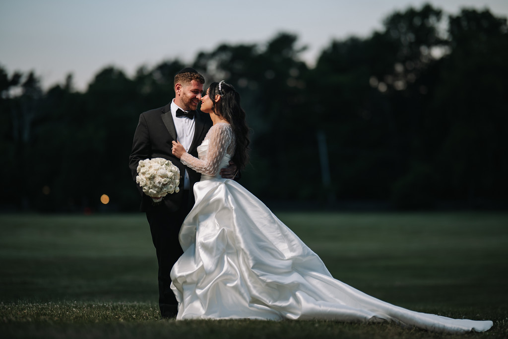 The bride in a flowing white gown and veil and the groom in a black suit share a kiss outdoors on the grass during golden-hour light.