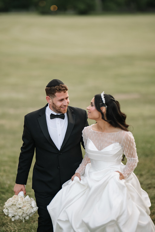 The Jewish bride in a white gown and veil and the groom in a black tuxedo stand on a grassy field holding hands and smiling at each other during the wedding ceremony.