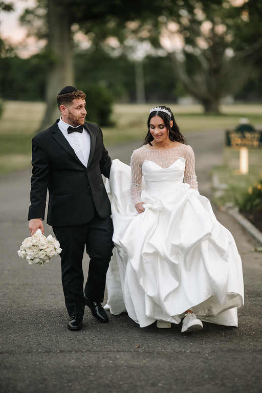 Frum bride in a white lace veil and ornate headpiece and groom in a black suit walk together outdoors on a tree-lined path during the wedding day.