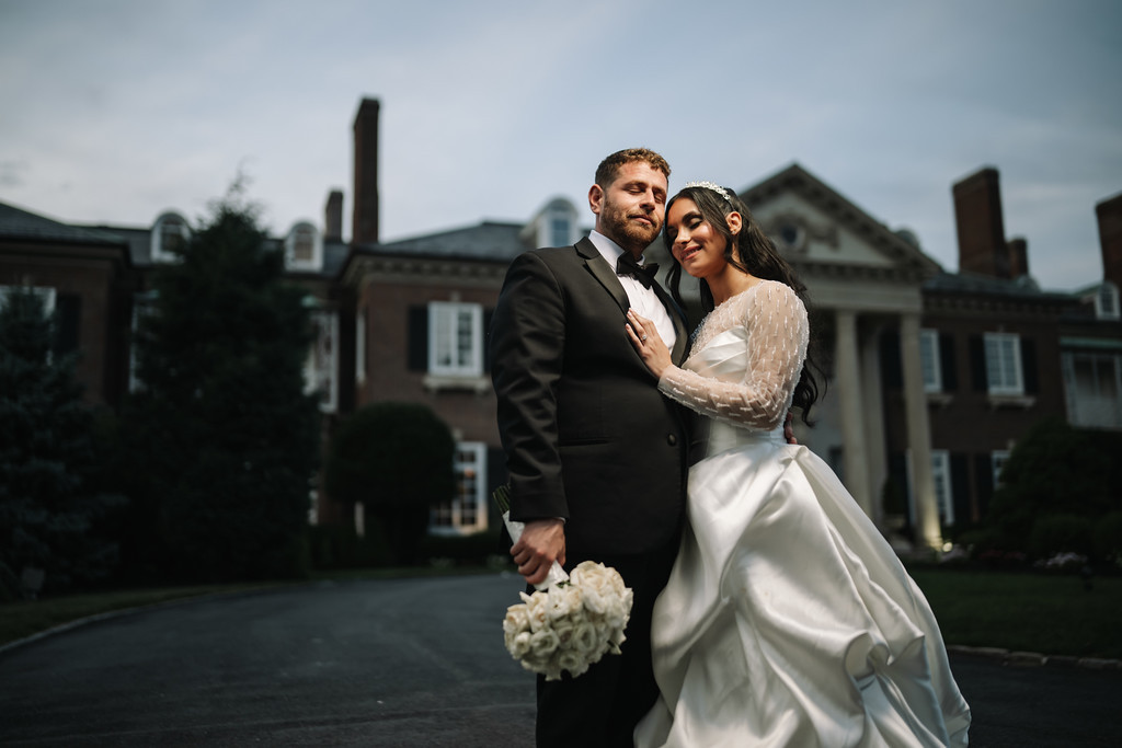 Newlywed couple posing in an embrace outdoors, bride in a white gown holding a bouquet while wearing a veil and hair adornment.