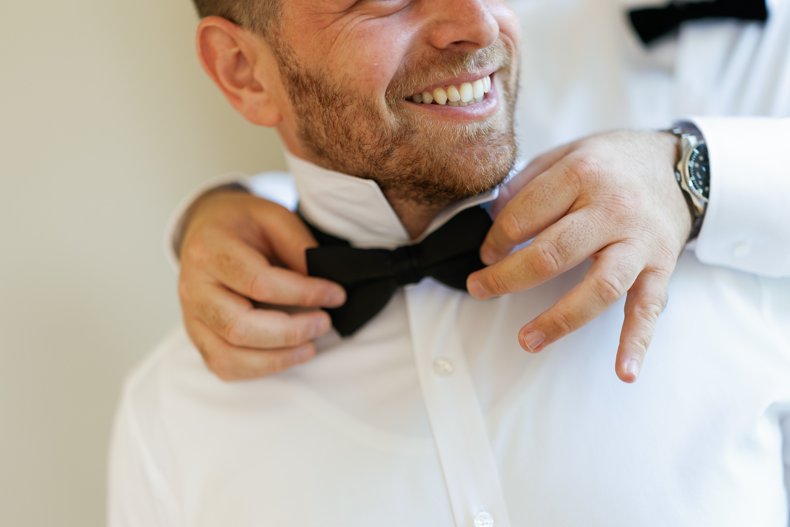 Smiling groom adjusting his bow tie while buttoning his white shirt at The Mansion At Glen Cove, photographed by CJ Studios