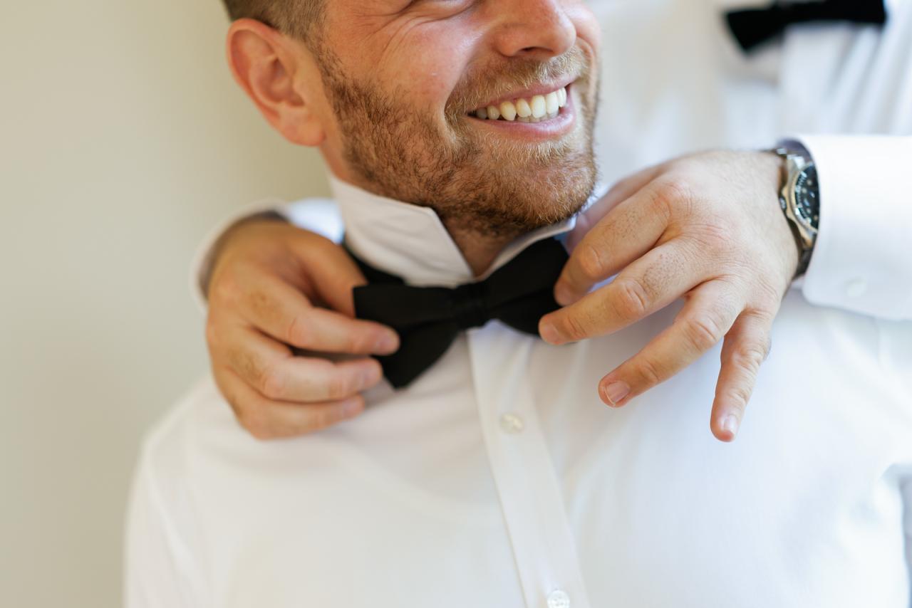 Smiling groom adjusting his bow tie while buttoning his white shirt at The Mansion At Glen Cove, photographed by CJ Studios. Wedding photography.